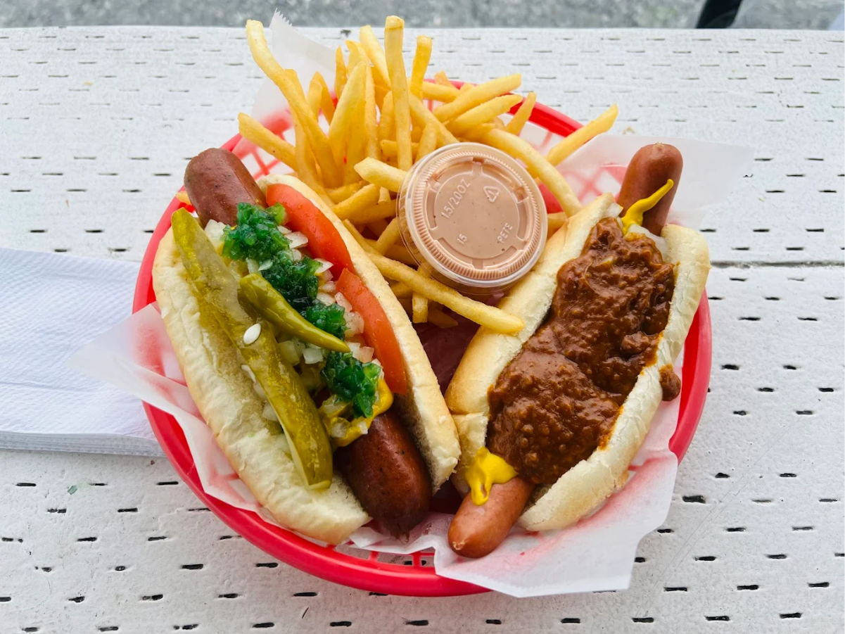 A red basket with two hot dogs—one topped with relish, tomatoes, onions, and peppers, the other with chili and mustard—served with a portion of French fries and a cup of dipping sauce on a white perforated table.