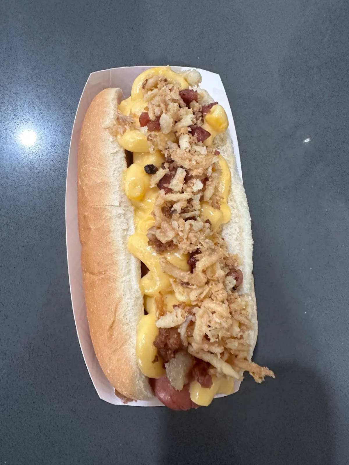 A hot dog in a soft bun topped with creamy macaroni and cheese, crispy fried onions, and bits of bacon, served in a white paper tray on a gray surface.