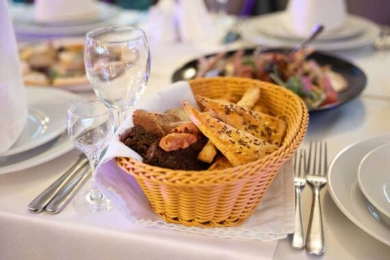 A wicker basket filled with assorted breads&mdash;classic steakhouse freebies&mdash;sits on a white tablecloth, surrounded by empty wine glasses, plates, and cutlery set for a meal. A salad is visible in the background.