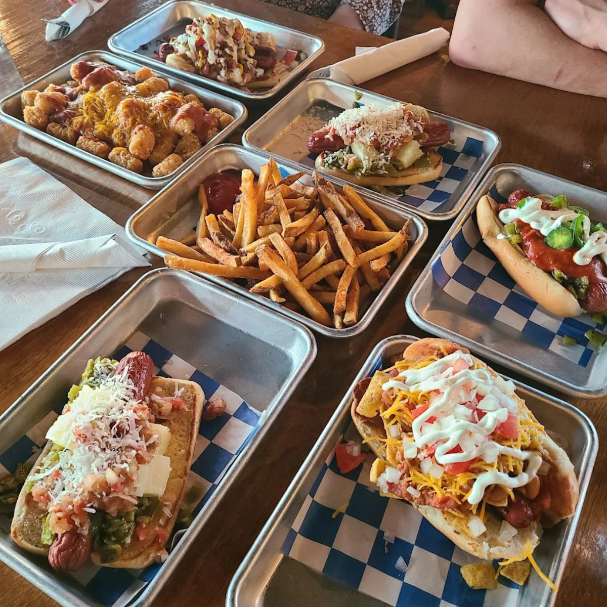 A table with trays of gourmet hot dogs topped with various ingredients, French fries, and tater tots on blue checkered paper, with a person's arms visible in the background.