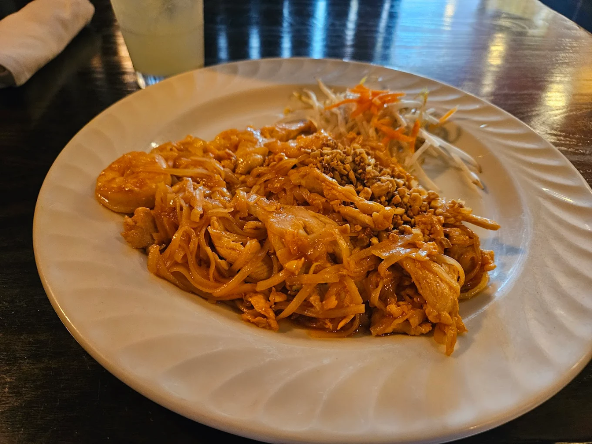 A plate of Pad Thai with shrimp, chicken, crushed peanuts, and a side of shredded carrots and cabbage, served on a white plate on a dark wooden table. A drink is partially visible in the background.