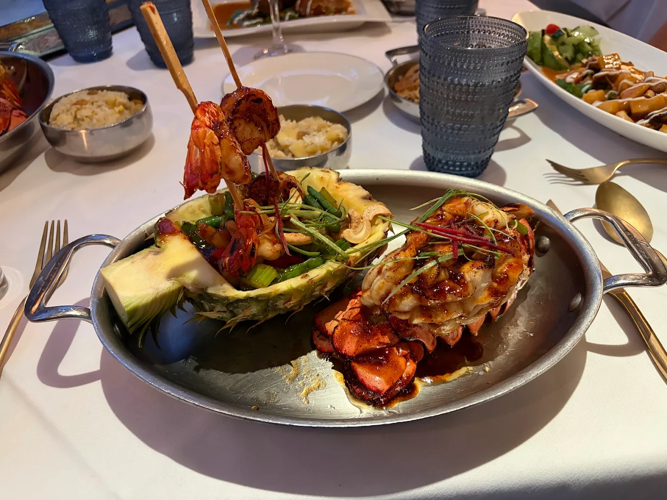 A metal serving dish with grilled lobster tail and shrimp skewers, garnished with herbs and served in a pineapple half with vegetables. Other dishes and drinks are on a white tablecloth in the background.