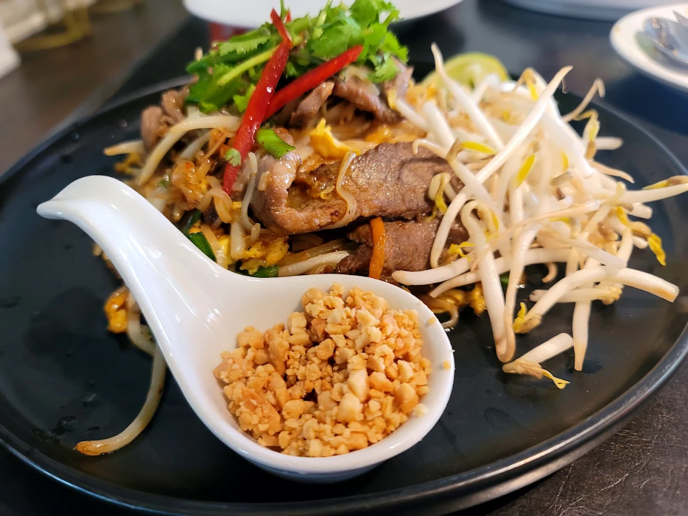 A black plate with stir-fried noodles, sliced beef, bean sprouts, and fresh herbs, garnished with red chili and cilantro. A white spoon holds crushed peanuts on the side.