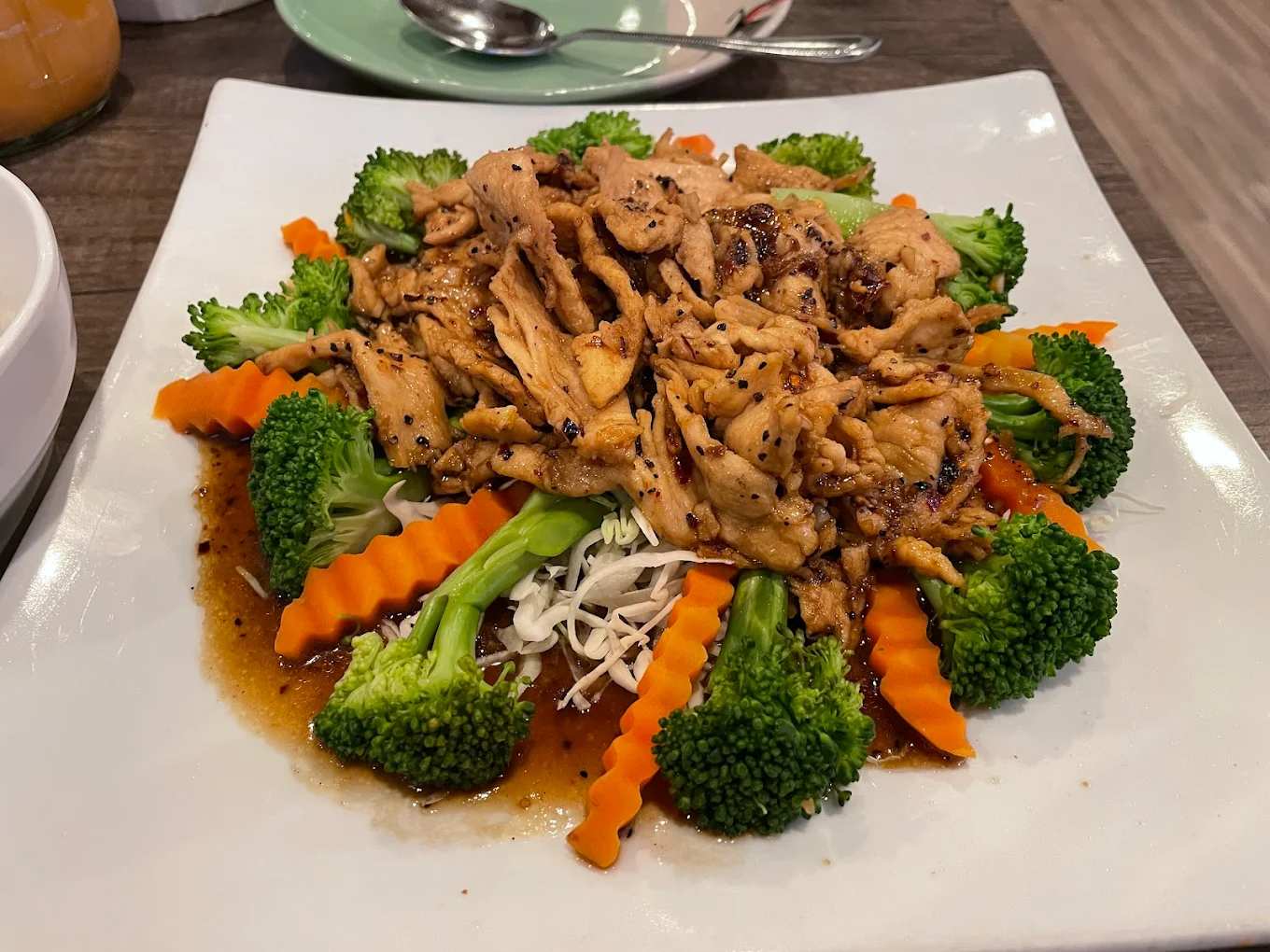 A white square plate with stir-fried sliced chicken in a brown sauce, surrounded by steamed broccoli florets and crinkle-cut carrot slices, served on a wooden table.
