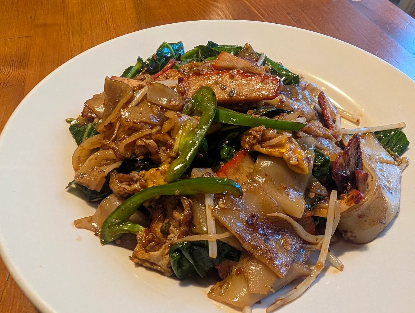 A white plate holds a serving of stir-fried wide rice noodles with vegetables, green peppers, leafy greens, bean sprouts, egg, and slices of meat, all on a wooden table.