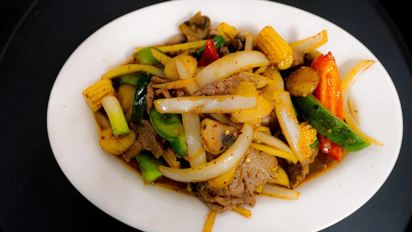 A white oval plate with stir-fried beef, onions, bell peppers, baby corn, and other vegetables in a savory brown sauce, set against a dark background.