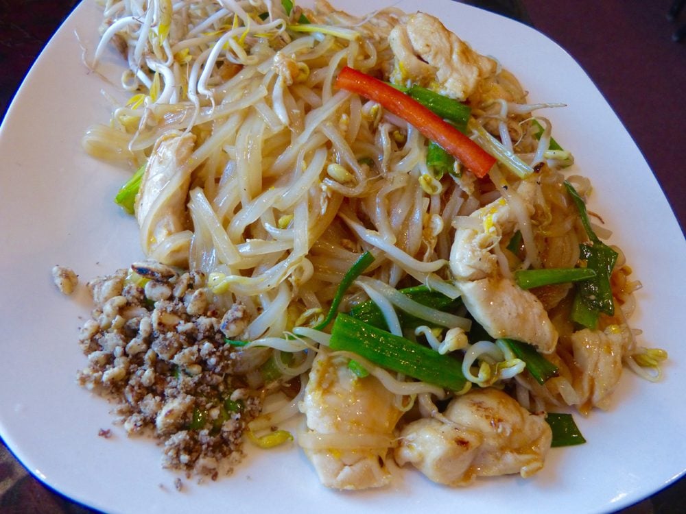 A plate of Pad Thai with rice noodles, bean sprouts, green onions, chicken pieces, a red chili strip, and a side of crushed peanuts on the corner of the white plate.