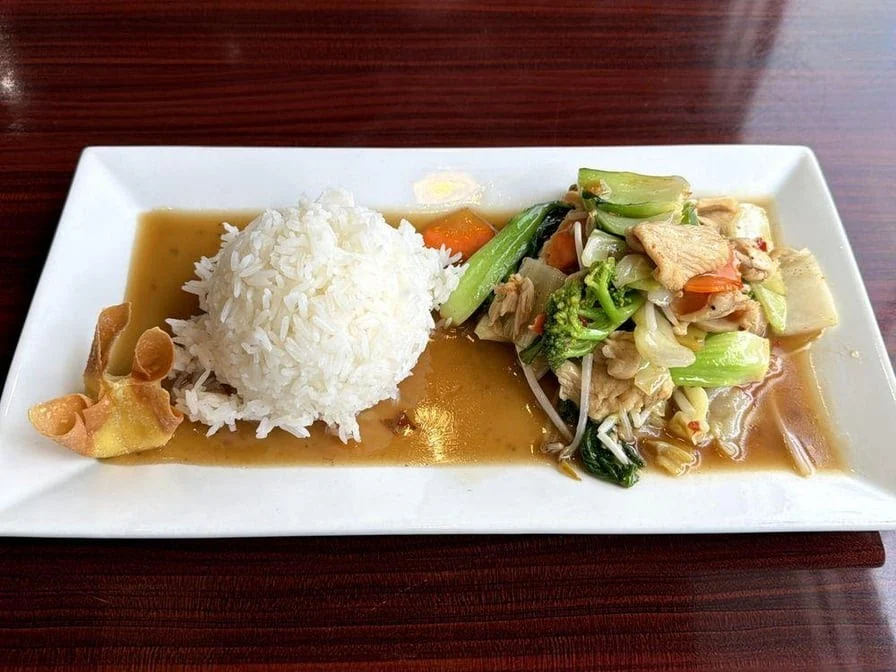A rectangular white plate with a serving of steamed white rice, mixed stir-fried vegetables, and meat in a brown sauce, garnished with a crispy chip, on a dark wooden table.