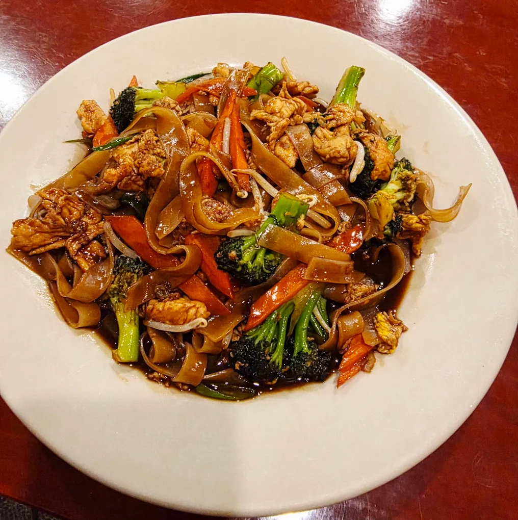 A white plate filled with stir-fried wide rice noodles, chicken, broccoli, carrots, and a dark savory sauce, on a reddish-brown tabletop.