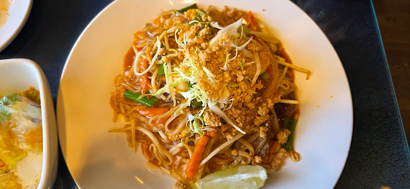 A plate of Pad Thai topped with crushed peanuts, bean sprouts, and vegetables, with a lime wedge on the side. The dish is served on a white plate.