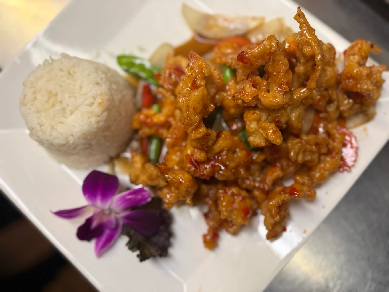 A plate of crispy fried chicken in a red sauce with vegetables, served with a mound of white rice and garnished with a purple orchid flower and leafy greens.