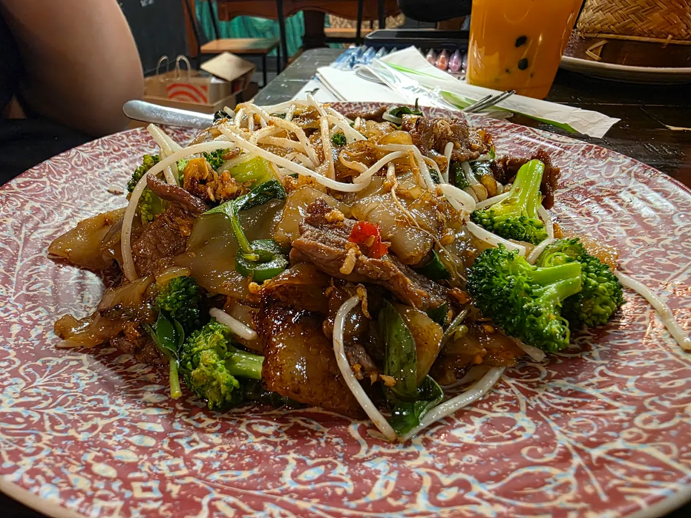 A plate of stir-fried noodles with beef, broccoli, bean sprouts, and vegetables, served on a patterned dish with a fork, with a drink and napkins in the background.