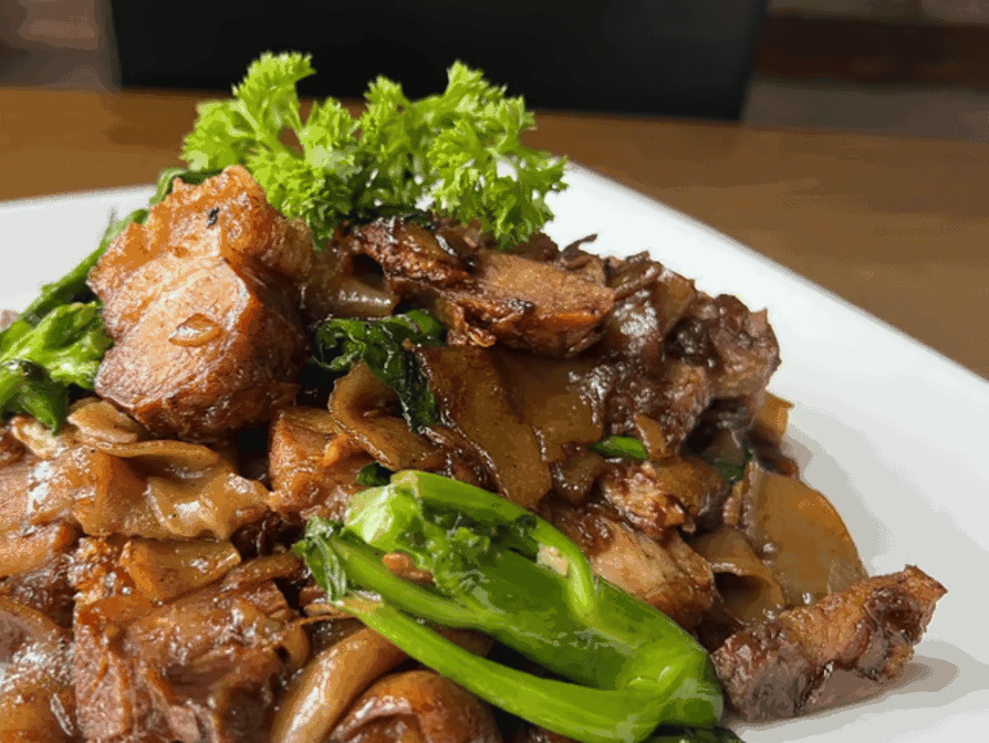 Stir-fried flat noodles with pieces of grilled meat and leafy green vegetables, garnished with fresh parsley, served on a white plate.