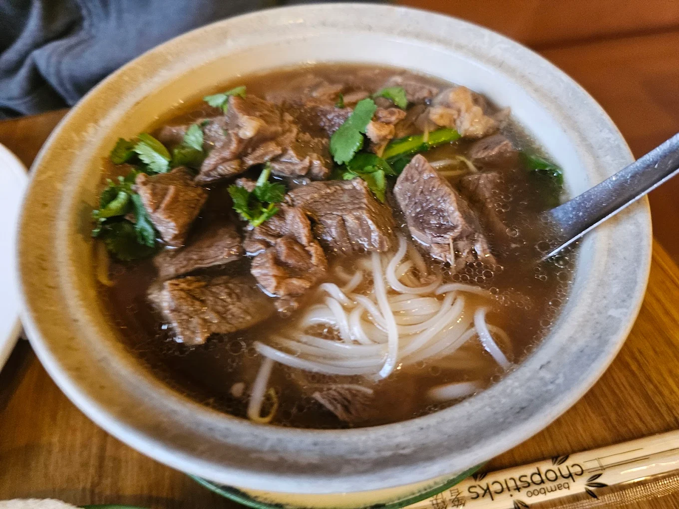 A bowl of beef pho with rice noodles, chunks of beef, fresh cilantro, and a spoon, served on a wooden table next to a pair of chopsticks.