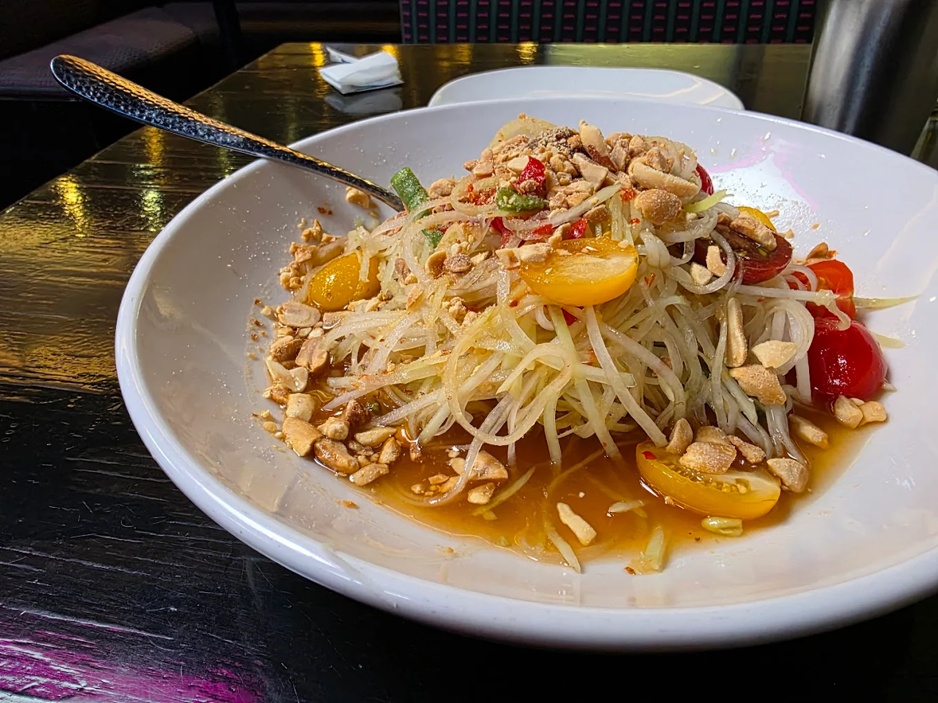 A white plate holds a serving of Thai green papaya salad topped with chopped peanuts, cherry tomatoes, and sliced green beans, with a spoon on the side. The dish is set on a dark, reflective table.