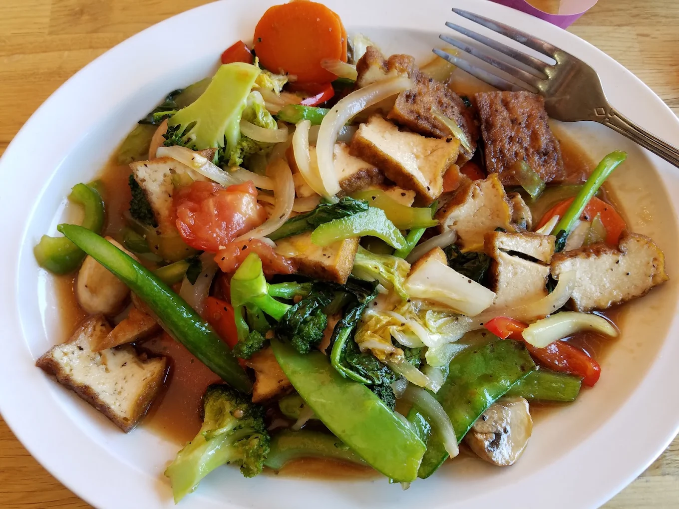 A white plate filled with stir-fried vegetables, tofu, broccoli, snow peas, onions, carrots, and tomatoes in a light brown sauce, with a fork on the plate.