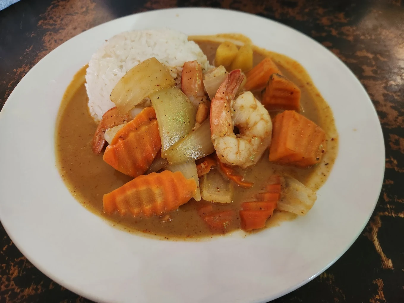 A white plate with rice and a yellow curry featuring shrimp, sliced carrots, onions, and potatoes, served on a dark, rustic table.