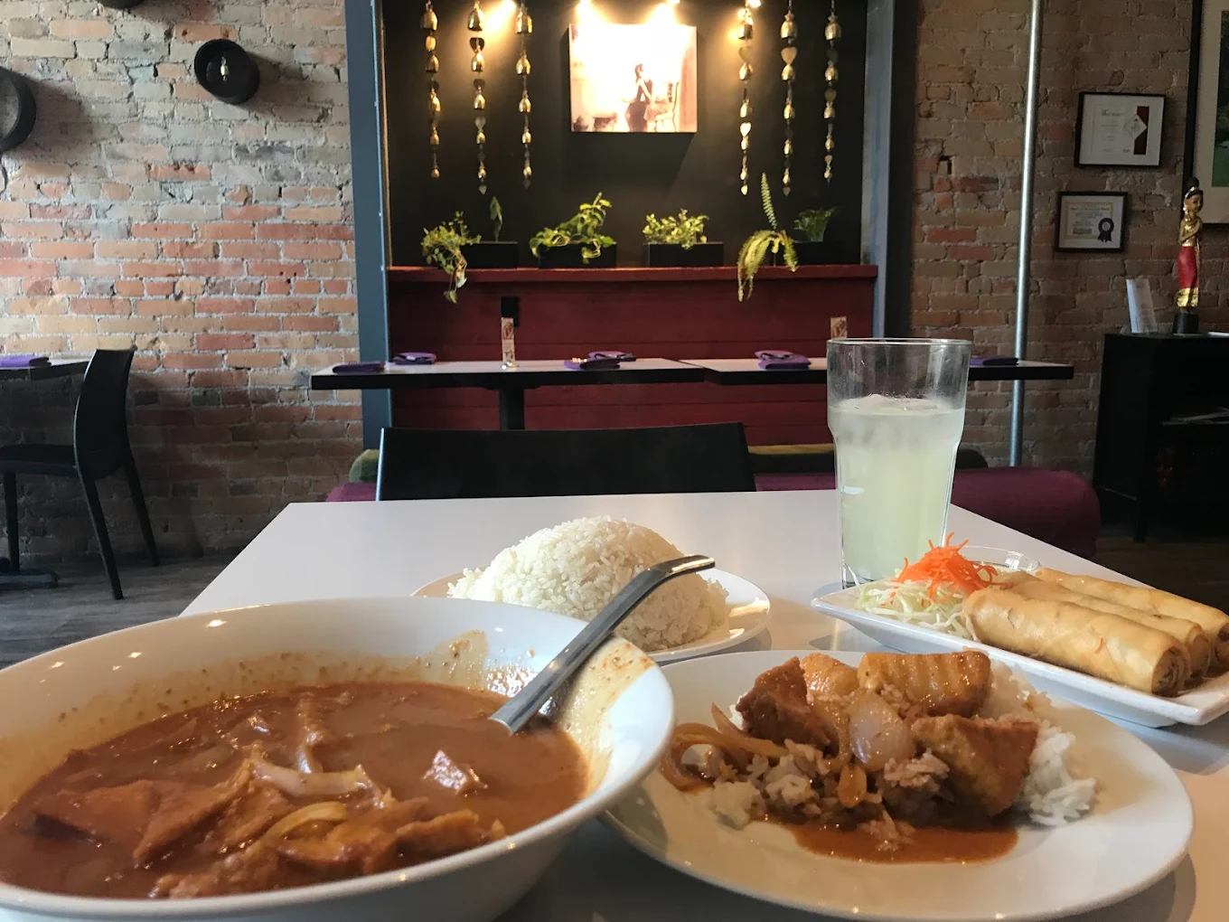 A table in a restaurant with a bowl of curry soup, a plate of white rice, fried spring rolls, and a glass of lemonade. The background features a brick wall, plants, and ambient lighting.