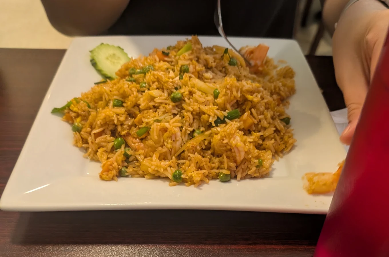 A person eating a plate of fried rice garnished with cut vegetables, using a fork. The dish is served on a square white plate, placed on a dark brown table. A cucumber slice is visible on the plate.