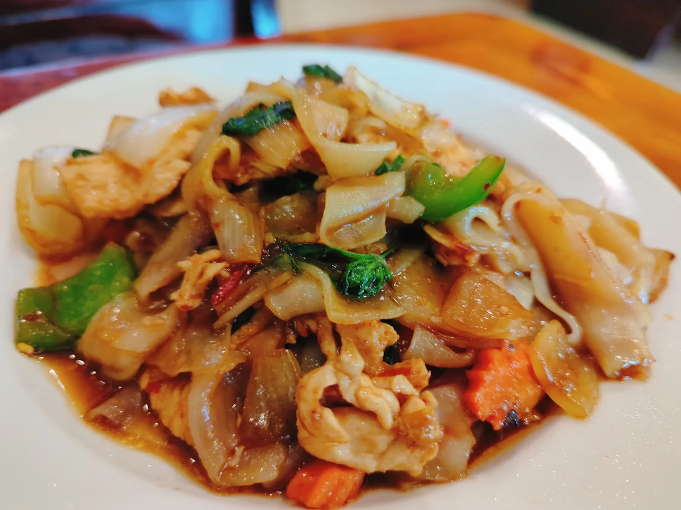 A close-up of a plate of stir-fried noodles with vegetables, chicken, and sauce, served on a white dish. The noodles appear glossy and are mixed with green peppers and leafy greens.