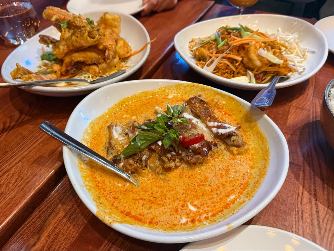 A table with plates of Thai food, including a bowl of red curry with vegetables and herbs, a dish of fried tempura, and a plate of noodles topped with shredded vegetables.