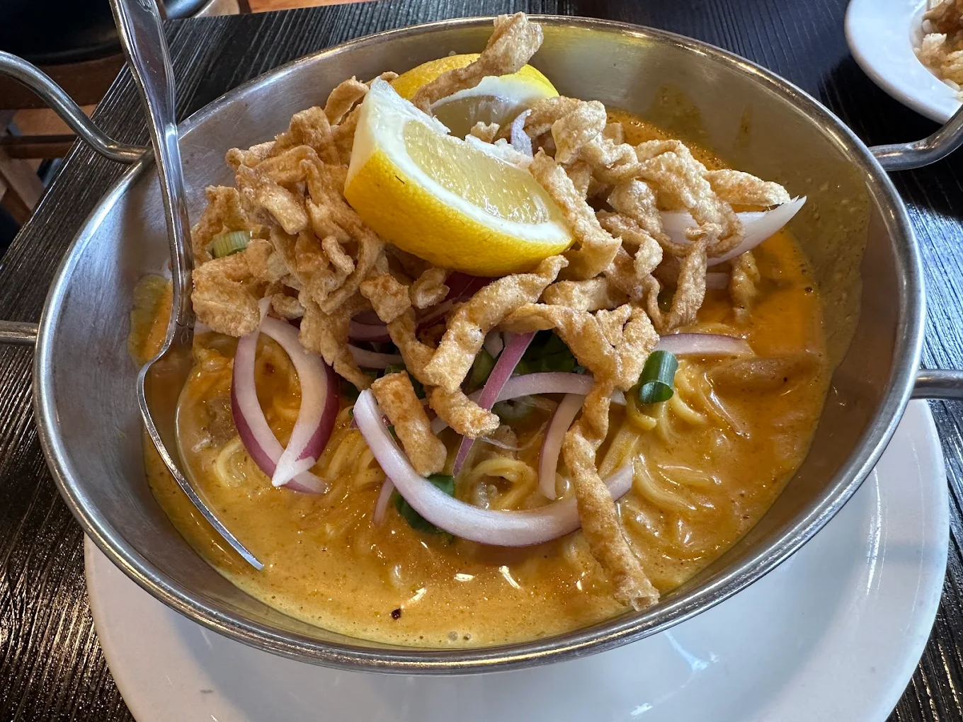 A bowl of creamy Thai curry noodle soup topped with crispy noodles, red onion slices, green onions, and two lemon wedges, served in a metal bowl with a spoon on the side.