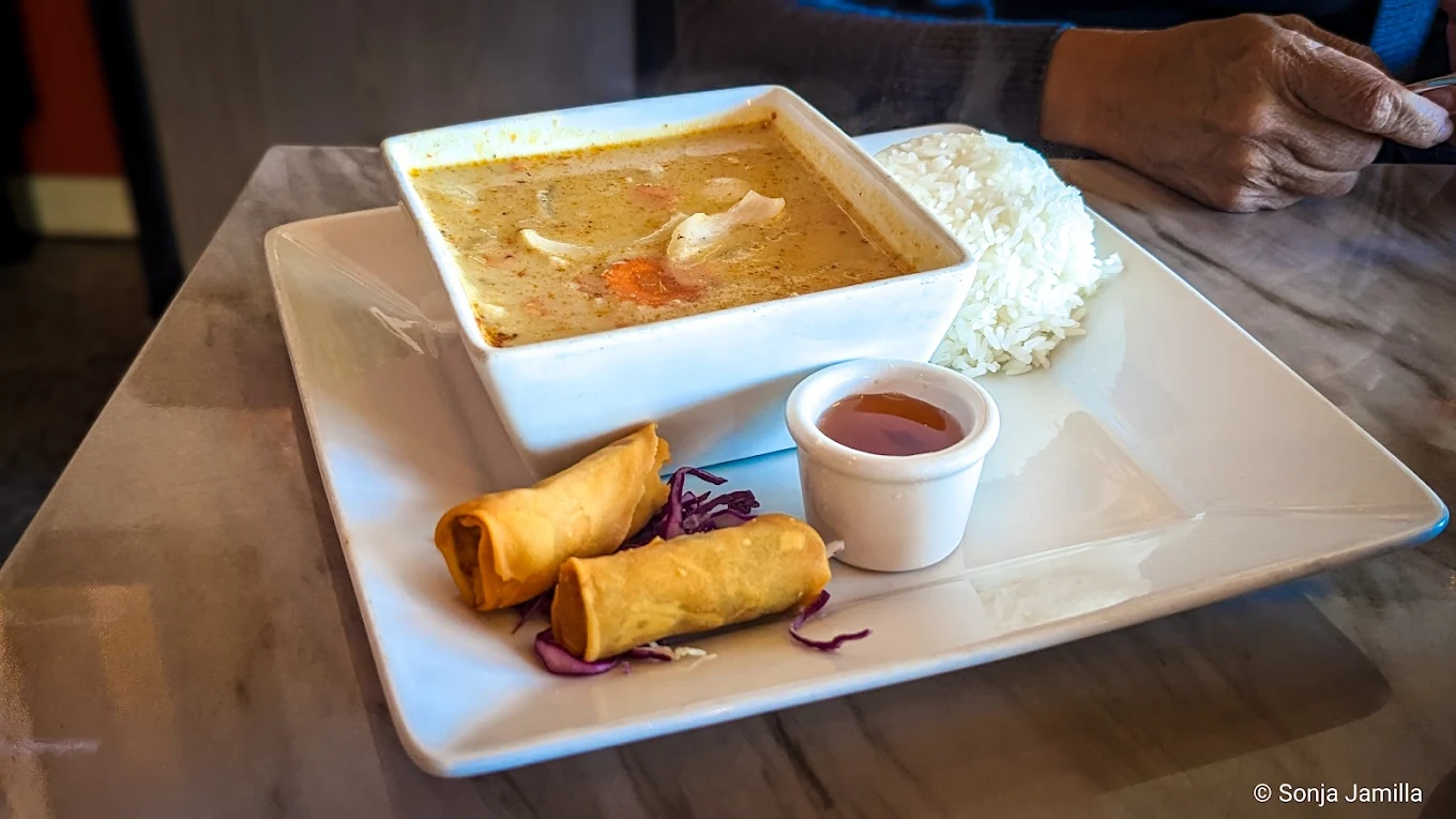 A square bowl of creamy soup with vegetables served alongside a mound of white rice, two spring rolls on shredded cabbage, and a small cup of dipping sauce on a white plate. A person's hand is visible in the background.