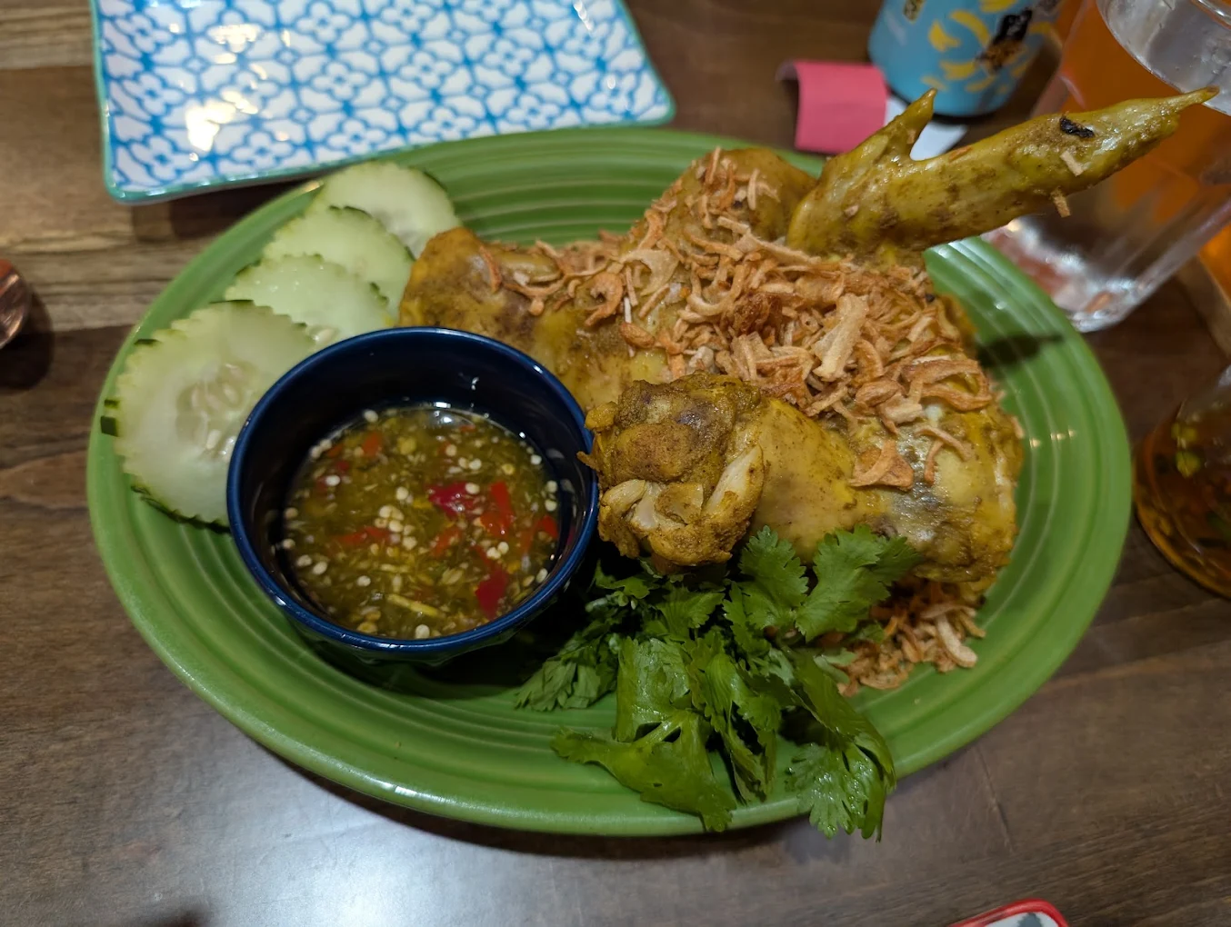 A green plate with fried chicken covered in crispy shallots, cucumber slices, cilantro, and a small bowl of spicy dipping sauce with chili and sesame seeds. A patterned dish is partially visible in the background.