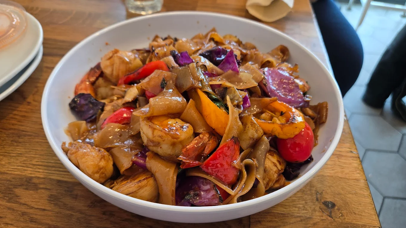 A white bowl filled with stir-fried wide rice noodles, shrimp, chicken, red and yellow bell peppers, purple cabbage, and vegetables in a brown sauce, placed on a wooden table.