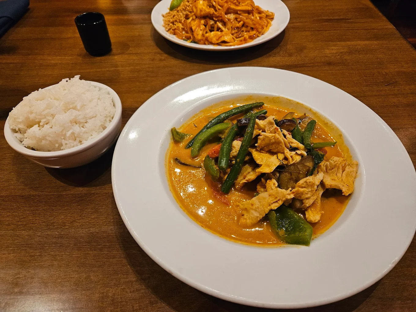 A plate of Thai curry with chicken, green beans, and bell peppers in orange sauce, served with a bowl of white rice. Another plate of noodles is visible in the background, all on a wooden table.