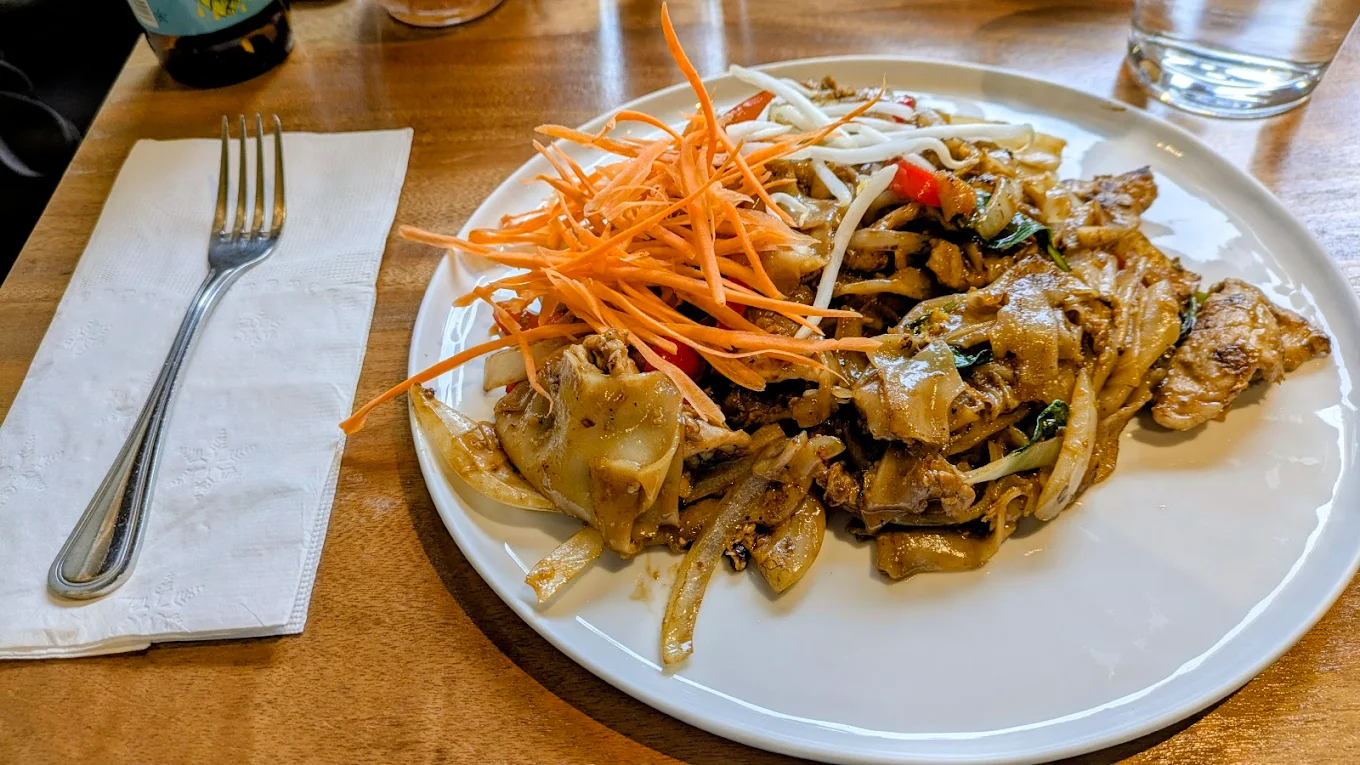 A white plate of stir-fried noodles with chicken, topped with shredded carrots and bean sprouts, sits on a wooden table next to a fork and napkin. A glass of water is visible in the background.