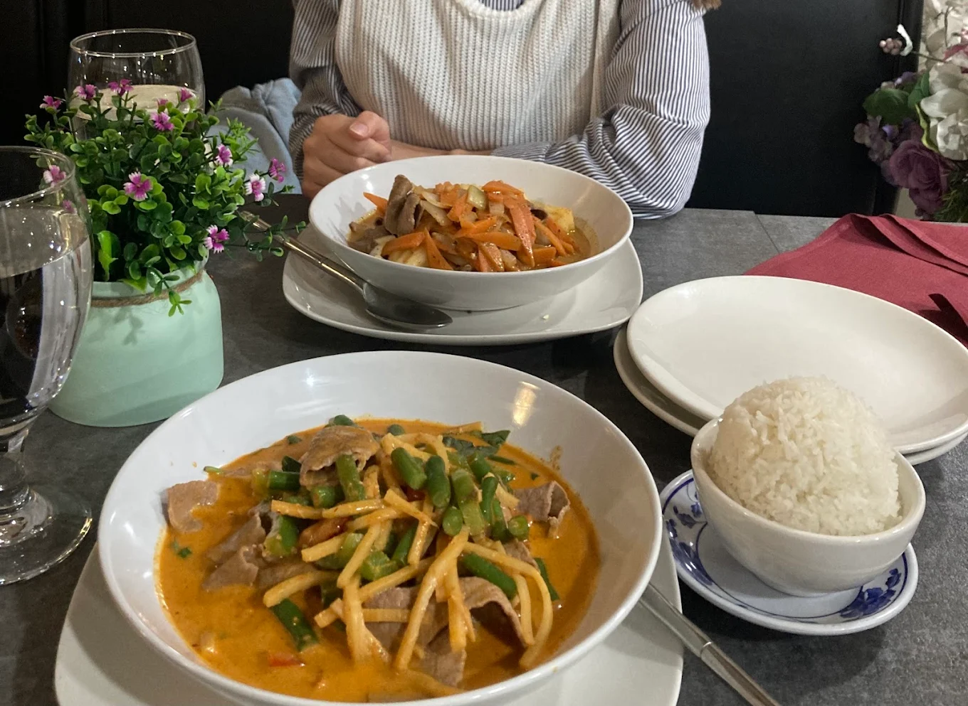 A table set with two plates of Thai curry dishes, a bowl of steamed white rice, glasses of water, and a small potted plant. A person sits across the table, hands folded near their chest.