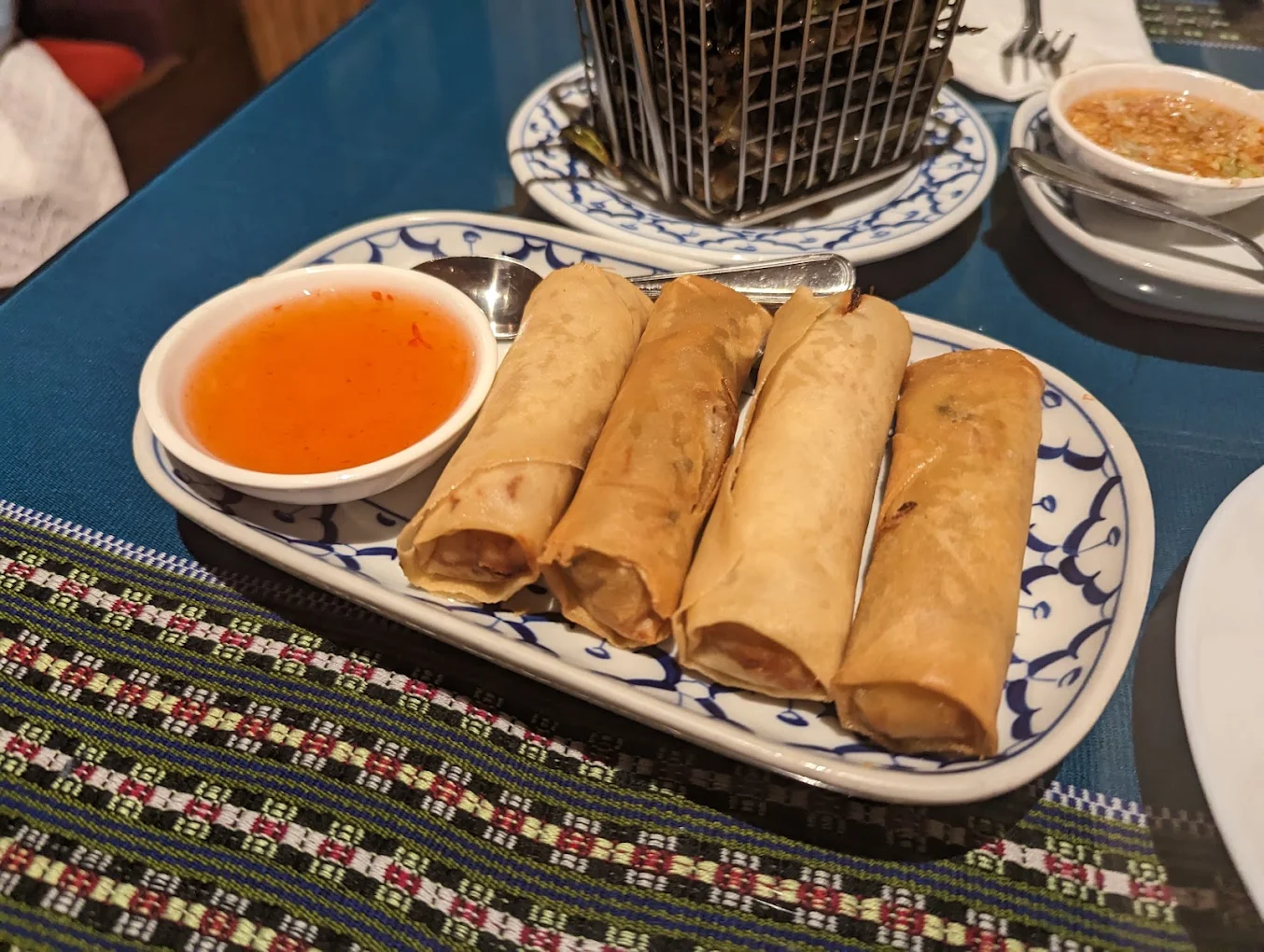 Four golden-brown spring rolls are arranged on a patterned plate next to a small dish of orange dipping sauce, atop a blue tablecloth with a decorative woven runner.