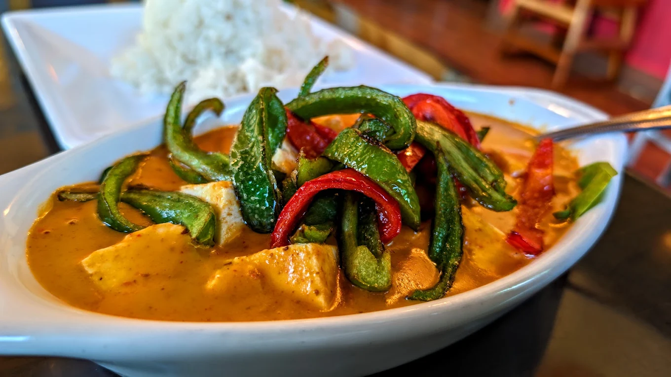 A close-up of a bowl of curry with colorful bell peppers and tofu in a creamy orange sauce, served with a side of white rice on a white plate in the background.
