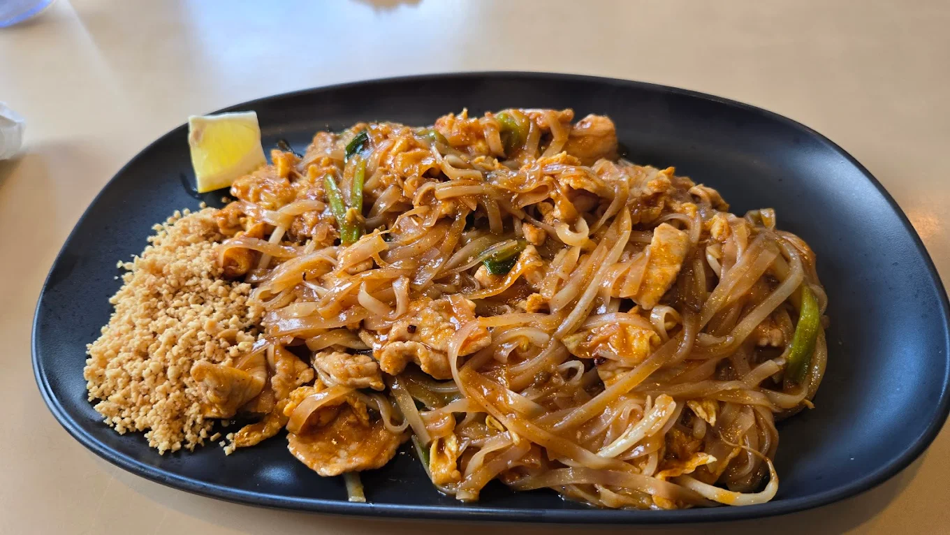 A black plate of Pad Thai with stir-fried noodles, chicken, egg, green onions, a wedge of lemon, and a side of crushed peanuts on a beige table.