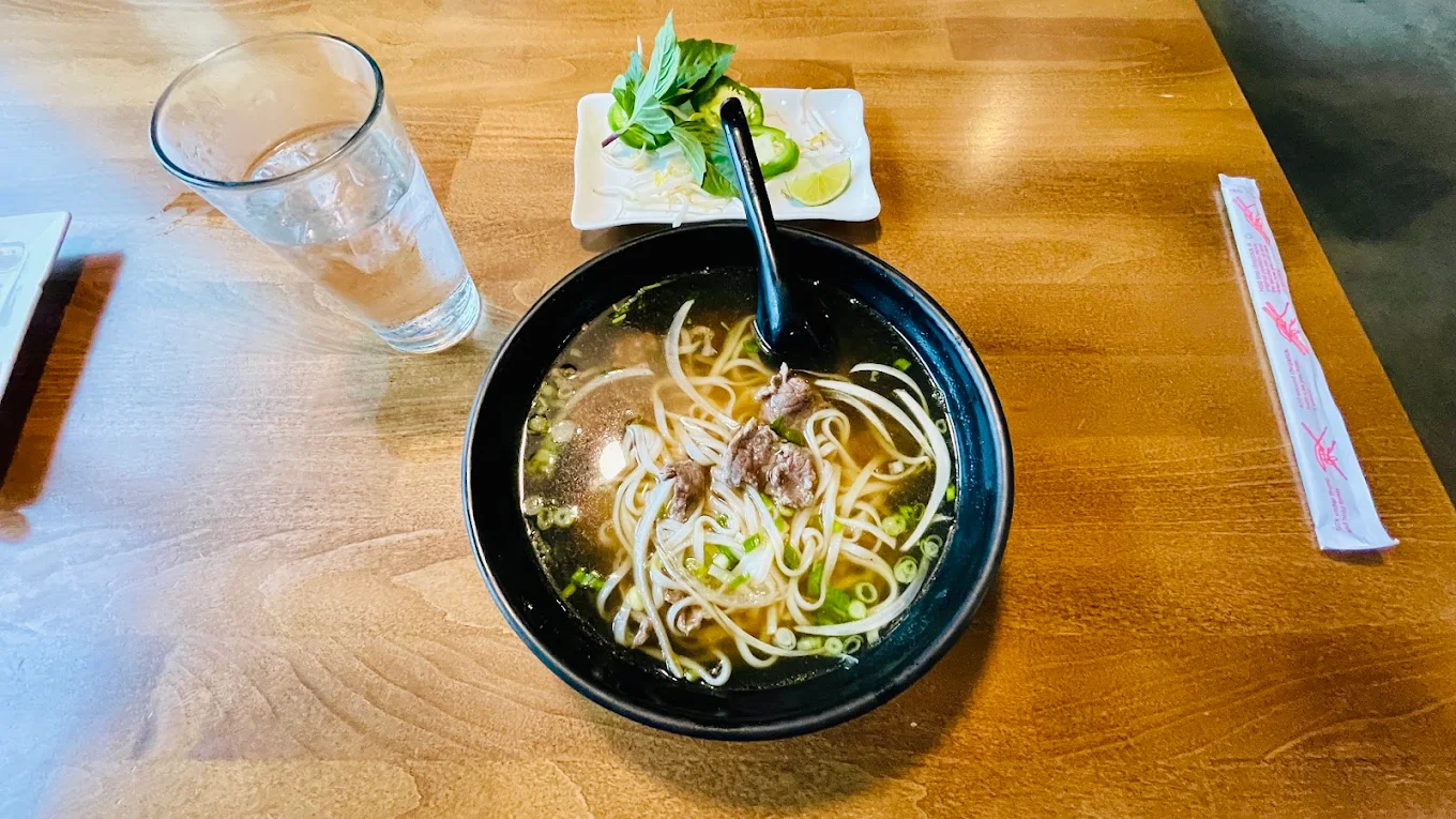 A bowl of beef pho with noodles, green onions, and broth sits on a wooden table. Nearby are a glass of water, chopsticks in a wrapper, and a plate with basil, lime, and bean sprouts.