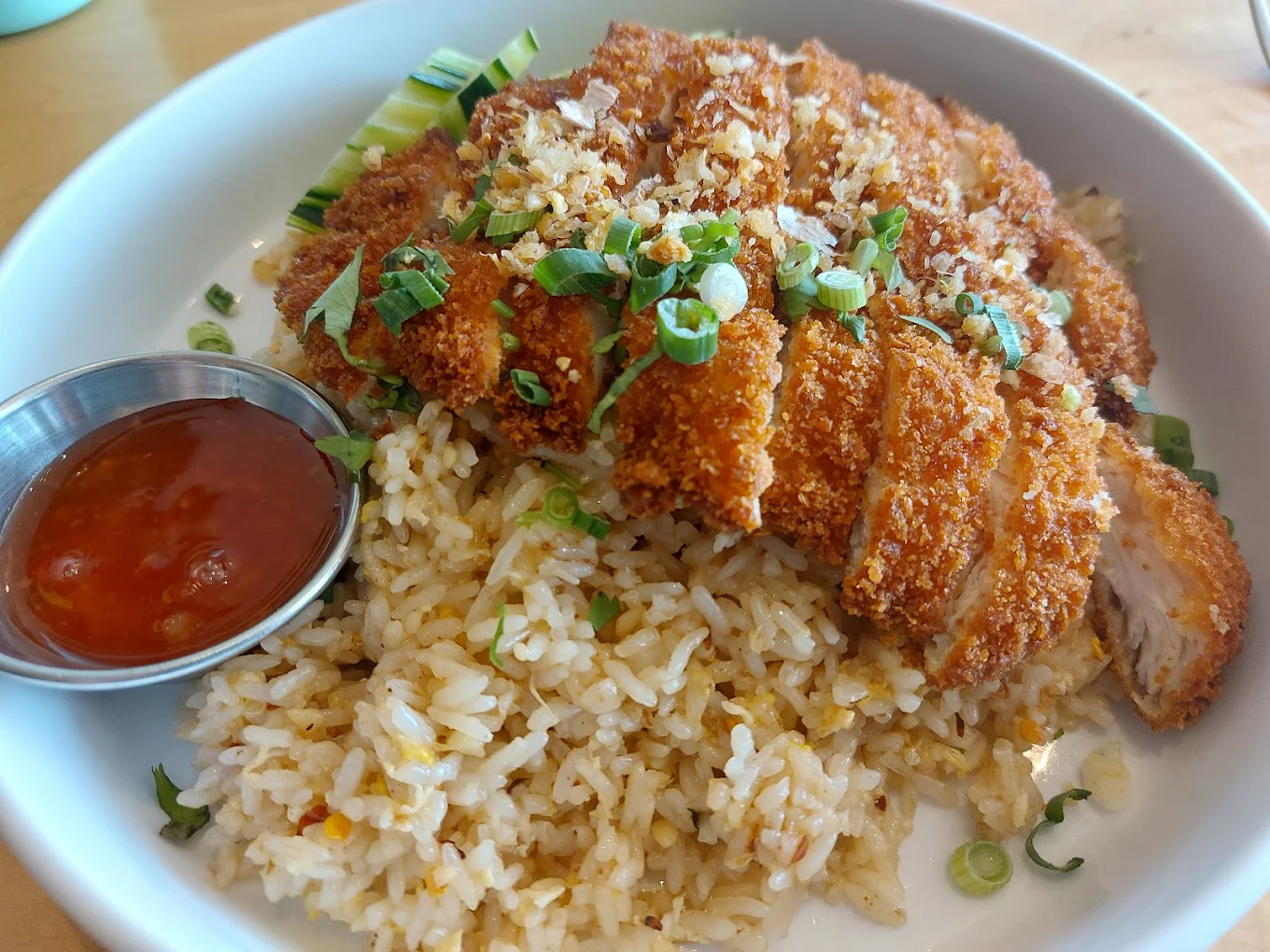 A white bowl filled with fried rice topped with sliced crispy breaded chicken, garnished with chopped green onions. A small metal cup of red dipping sauce is on the side. Slices of cucumber rest in the background.