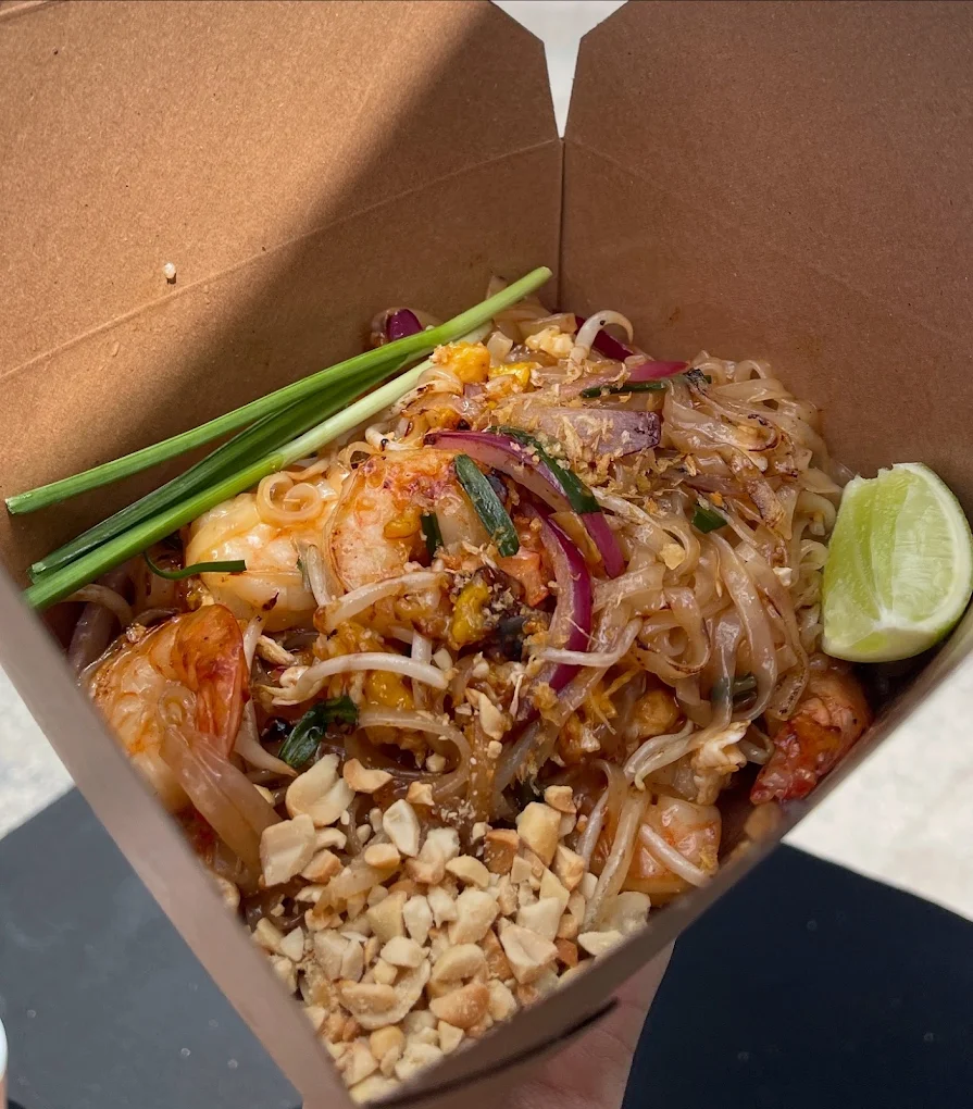 A takeout box of Pad Thai noodles topped with shrimp, crushed peanuts, red onions, green onions, and a lime wedge, served outdoors in natural light.