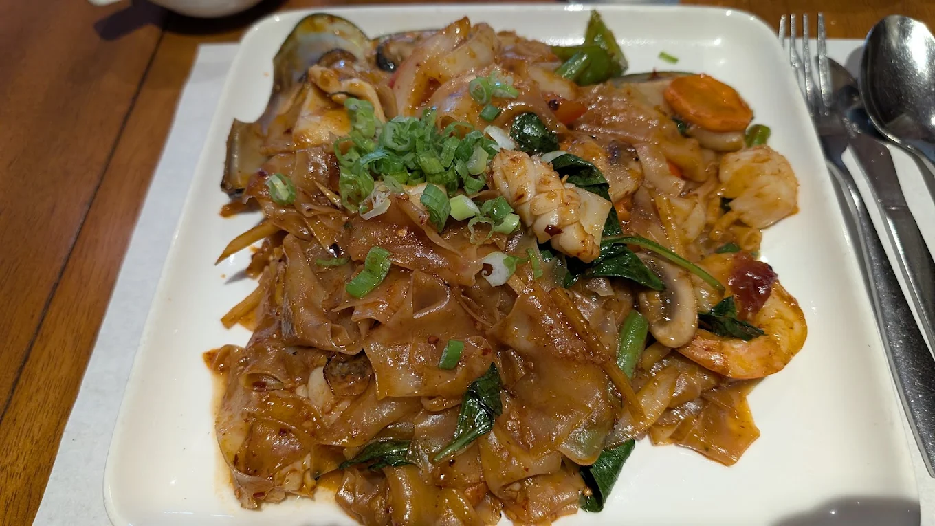 A square white plate filled with stir-fried flat rice noodles, shrimp, squid, green vegetables, carrots, and mushrooms, garnished with chopped green onions. A fork and spoon are placed on the right side of the plate.