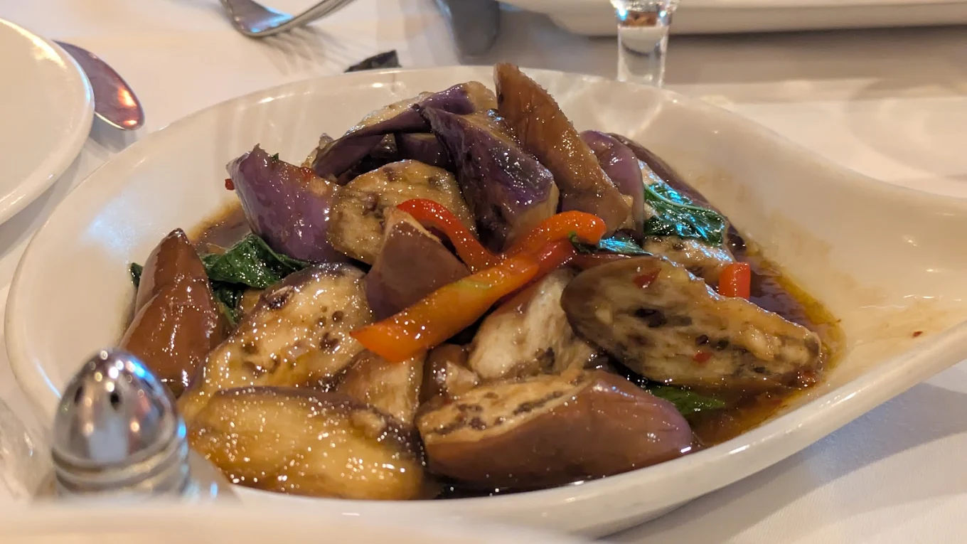 A white dish filled with stir-fried eggplant, bell peppers, and leafy greens in a savory brown sauce, served on a table set for a meal.