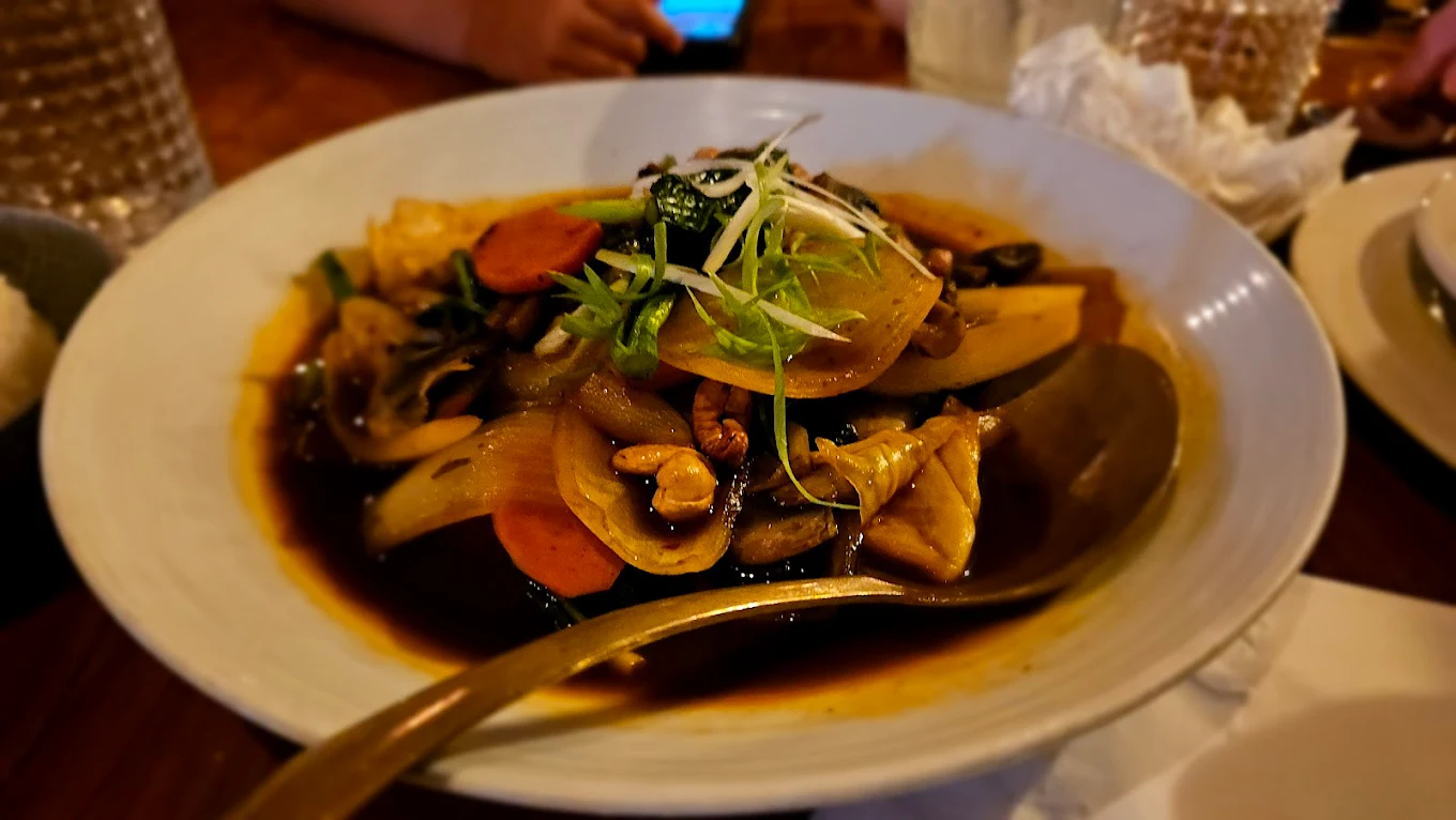 A bowl of stir-fried vegetables, sliced carrots, onions, and meat in a dark sauce, garnished with green onions, with a large serving spoon resting in the dish on a wooden table.