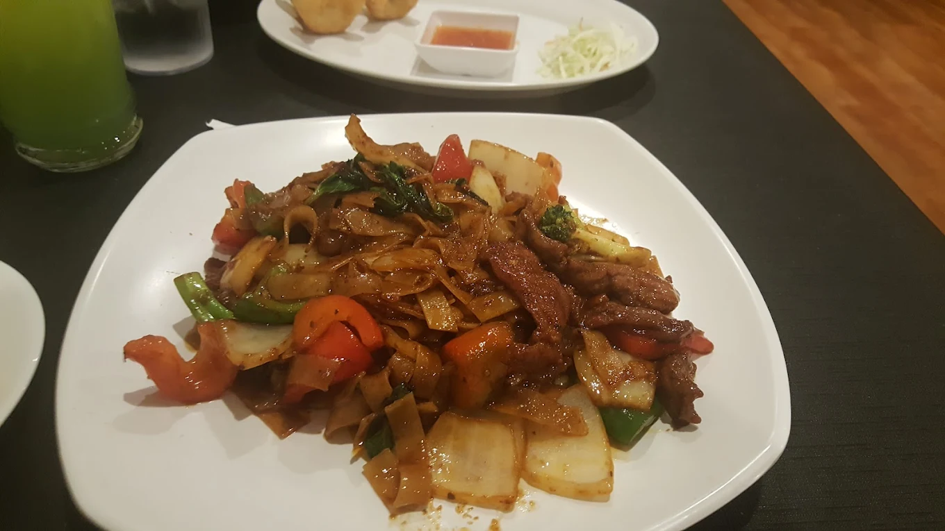 A plate of stir-fried flat noodles with sliced beef, bell peppers, onions, and other vegetables sits on a white dish on a dark table. A drink and another plate with appetizers and dipping sauce are in the background.