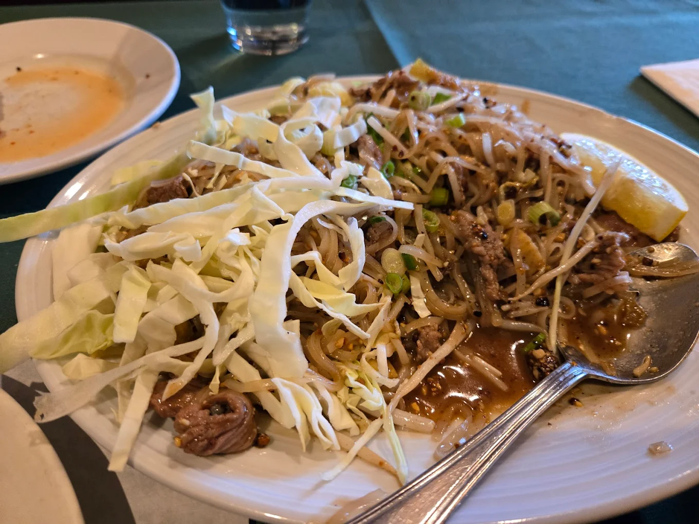 A plate of stir-fried noodles with beef, bean sprouts, and shredded cabbage, served with a spoon. A partially eaten appetizer on a small plate and a glass of water are in the background.
