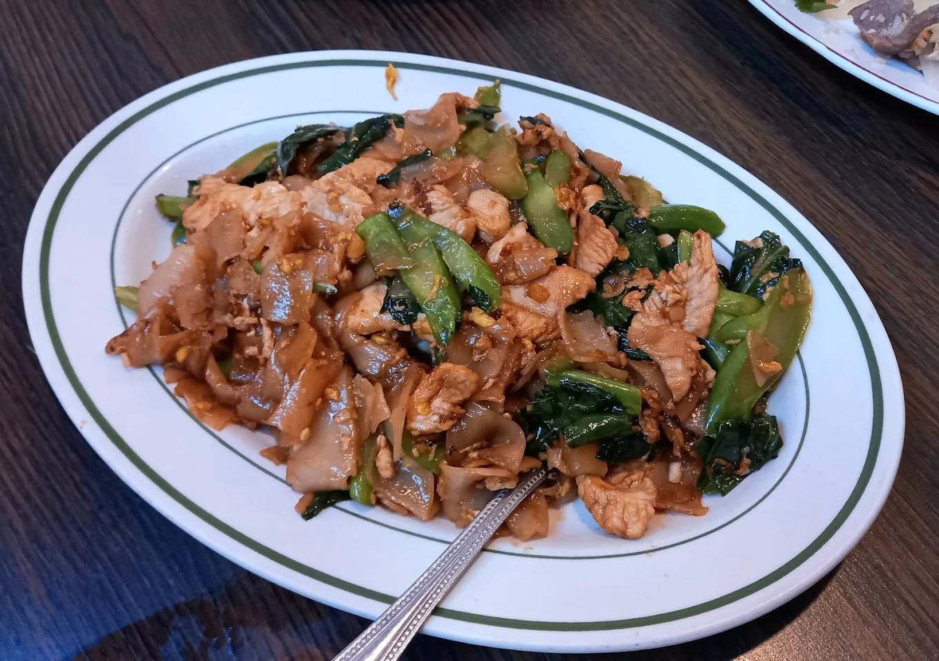 A plate of stir-fried flat rice noodles with pieces of chicken, Chinese broccoli, and egg, served on a white oval dish with a green rim, with a metal spoon resting in the food.