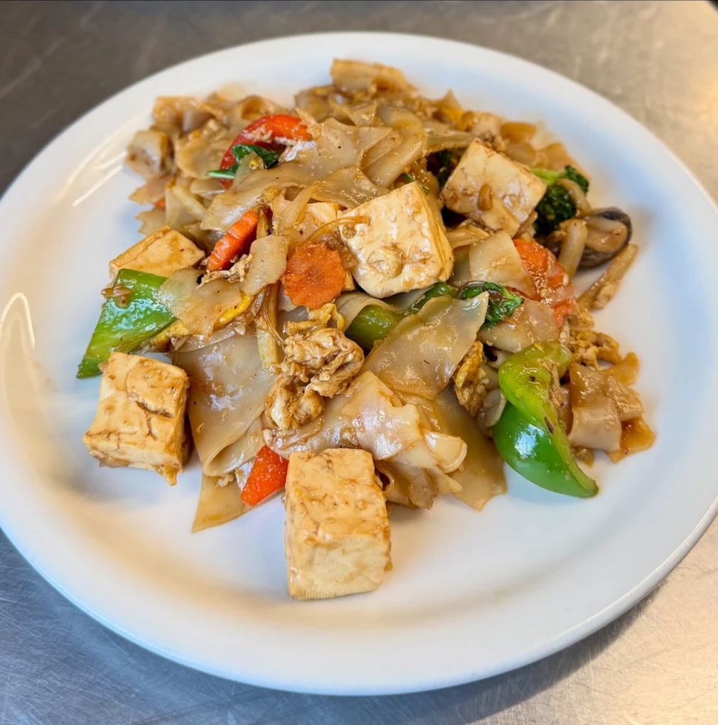 A white plate filled with stir-fried flat rice noodles, tofu cubes, broccoli, carrots, bell peppers, and mushrooms in a savory sauce.