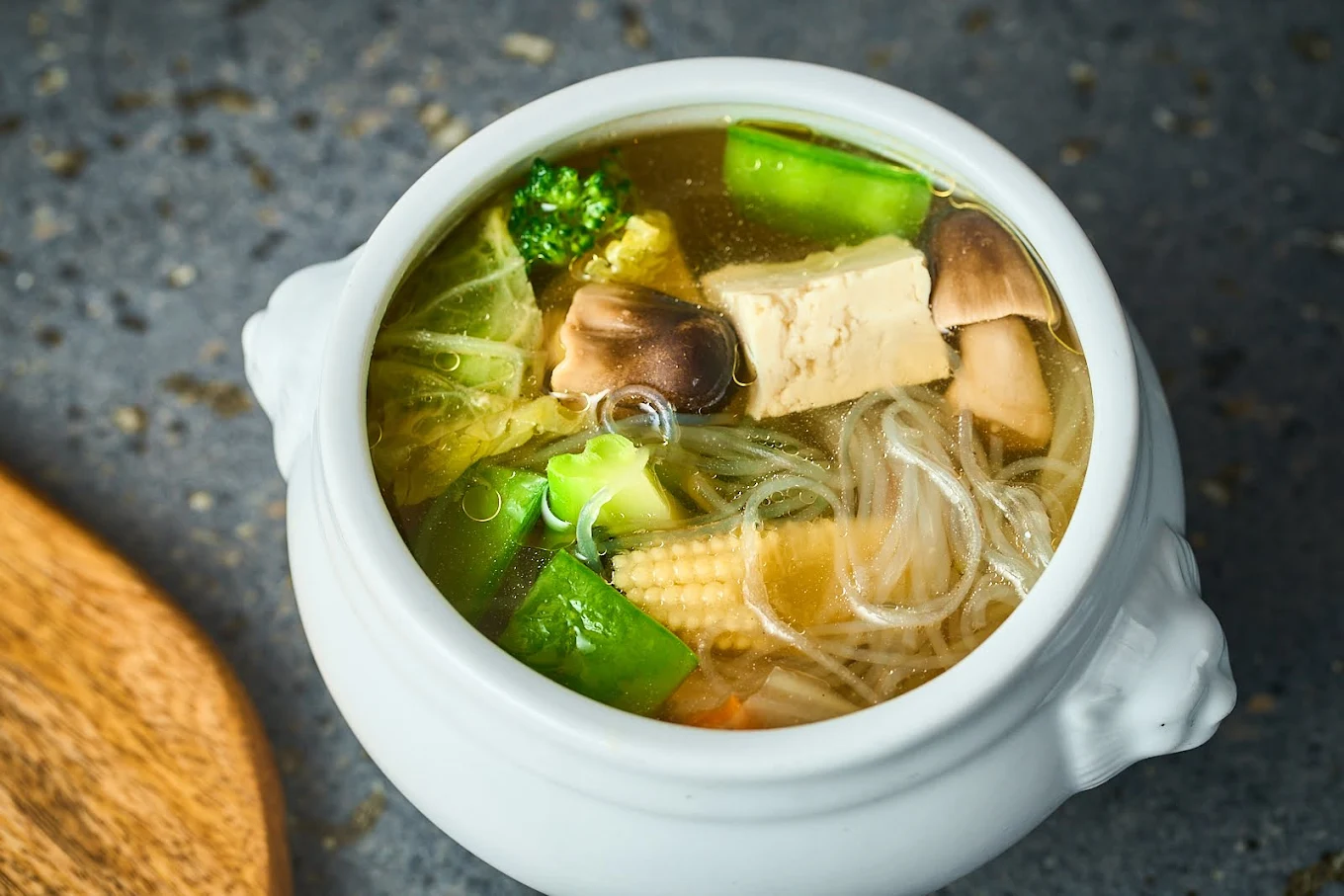 A white bowl filled with clear vegetable soup containing tofu, mushrooms, baby corn, broccoli, snow peas, cabbage, and glass noodles, placed on a gray surface.