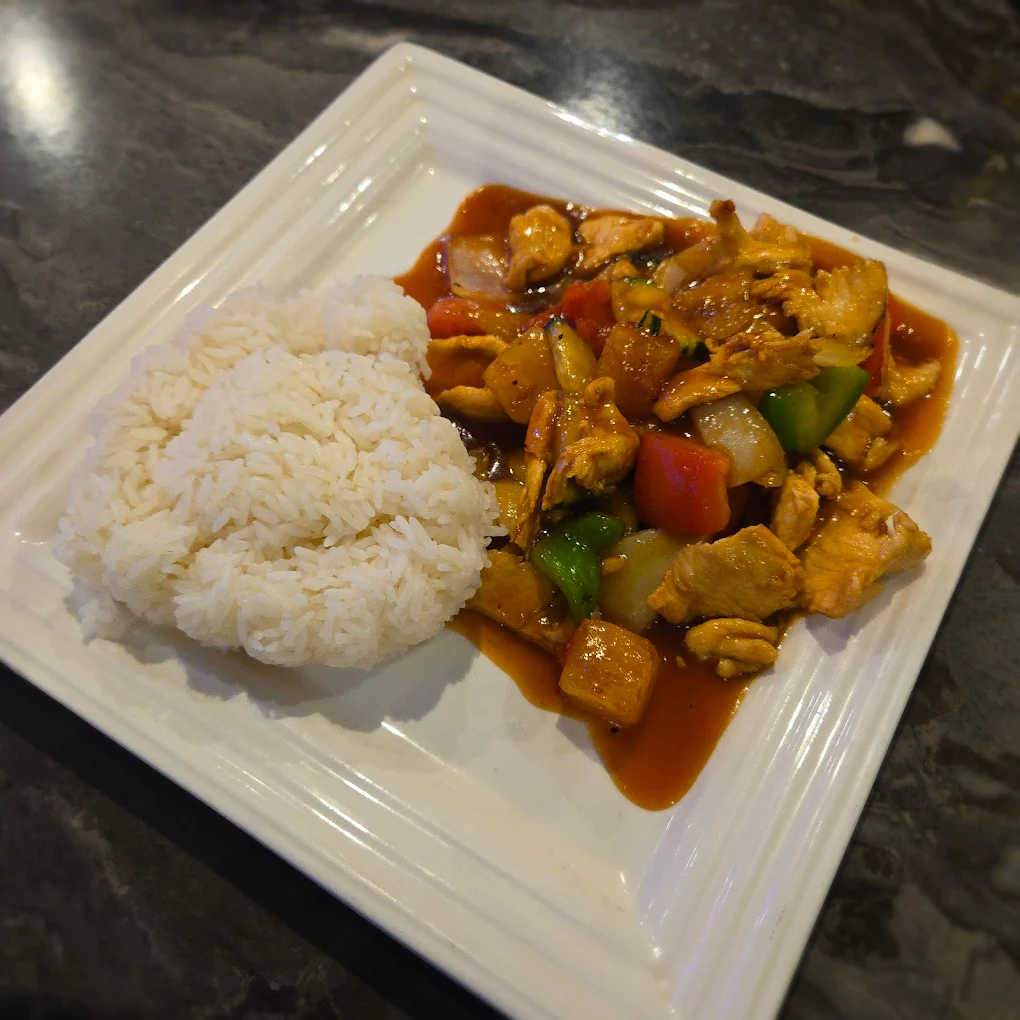 A square white plate with a serving of white rice shaped in a half-circle next to stir-fried chicken, bell peppers, and onions in brown sauce on a dark marble surface.
