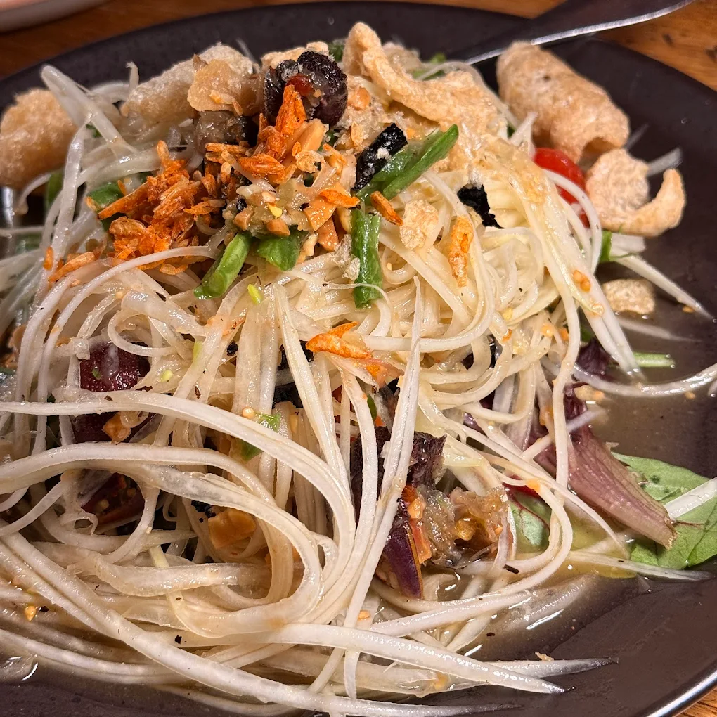 A close-up of Thai papaya salad (som tam) on a dark plate, featuring shredded green papaya, green beans, tomatoes, dried chilies, crispy pork rinds, and topped with fried garlic, all mixed in a light dressing.