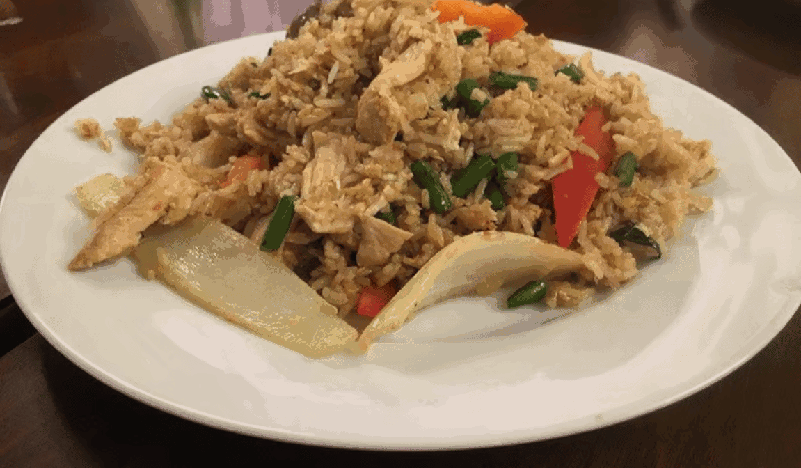 A plate of fried rice with pieces of chicken, sliced red bell pepper, green beans, and chunks of onion on a white dish, served on a dark wooden table.