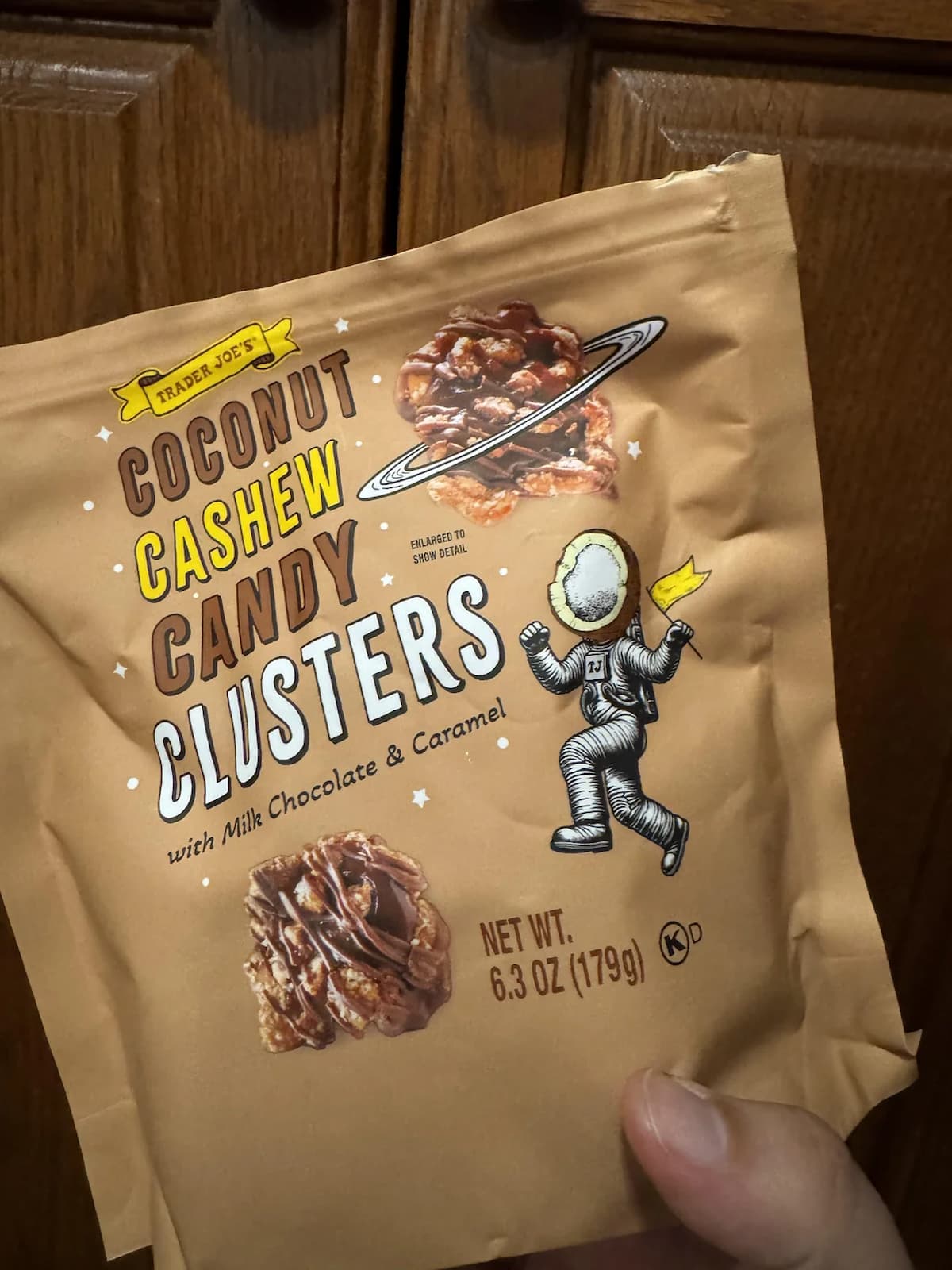 A hand holds a bag of Trader Joe's Coconut Cashew Candy Clusters with milk chocolate and caramel. The packaging features candy images and an astronaut illustration against a brown background.